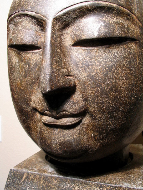 Sold!<br /><br />
DESCRIPTION: A large and finely sculpted head of the Buddha, the round face finely formed with serene downcast eyes, conveying a feeling of great gentleness and devotion. The crisp rounded brows meet at the narrow straight nose, ending in bud-like pursed lips which curl in an enigmatic smile.  The head is covered in tightly curled knots over pendulous ears.  Like the best of Chinese sculpture, this regal yet accessible representation of the Buddha successfully transforms Buddhist doctrine into inspirational shape. 
<p>The encrustations inside the carved lines, as well as the lightly mottled surface of the dark stone suggests an age earlier than the one given, especially when compared to the light scuff on one upper eyelid or the small chip in the right ear which are obviously more recent than the sculpture itself.  We have priced this as a 19th C. piece however, as we do not have confirmation of its age.  The head rests on a separate trapezoidal base.  DIMENSIONS: Including base, 21” high (53.5 cm) x 11” wide (28 cm) x 13 ½” deep (34 cm).  Shipping to be handled by Craters and Freighters.  
<div id='rater_target1089300'></div>
